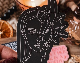 May include: Black and white line art sticker of a woman's face with a dragon's head emerging from her forehead. The sticker is on a wooden surface with other stickers and a candle.