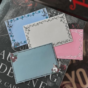 May include: Three decorative paper note cards with floral and barbed wire borders. The top card is light blue, the middle card is white, and the bottom card is light blue with a skull and crossbones design.