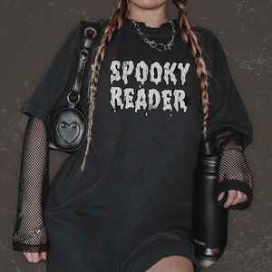 May include: Black oversized t-shirt with the text "Spooky Reader" in white dripping letters. The shirt is worn with black fishnet sleeves and a black heart-shaped bag.
