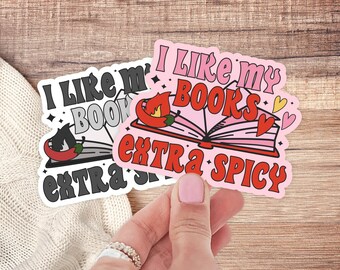 Pode incluir: Dois adesivos com o texto "Eu gosto dos meus livros extra picantes" e uma imagem de desenho animado de um livro com uma pimenta. Os adesivos são rosa e cinza com detalhes vermelhos e amarelos.