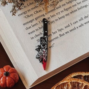 May include: A black and red knife-shaped enamel pin with a floral design and a blood-like effect. The pin is set against an open book with text. Decorative dried orange slices and a small orange pumpkin are also visible.