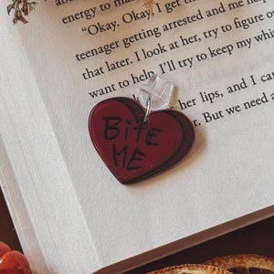 May include: A heart-shaped charm with the words "BITE ME" in black lettering. The charm is dark red with a darker outline and is attached to a clear pin. The charm is resting on the open pages of a book.
