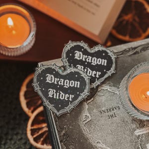 May include: Two black and white stickers with a barbed wire border and the text "Dragon Rider" in a gothic font. The stickers are placed on a brown leather-bound book with a map printed on the cover.