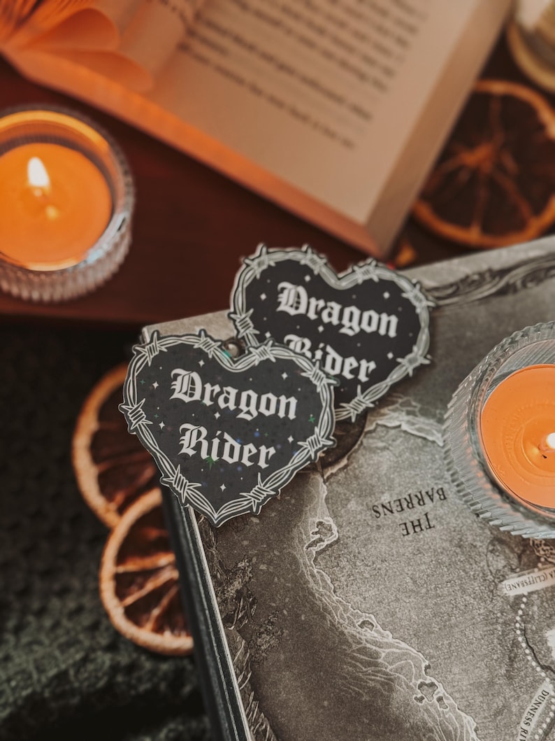 May include: Two black and white stickers with a barbed wire border and the text "Dragon Rider" in a gothic font. The stickers are placed on a brown leather-bound book with a map printed on the cover.