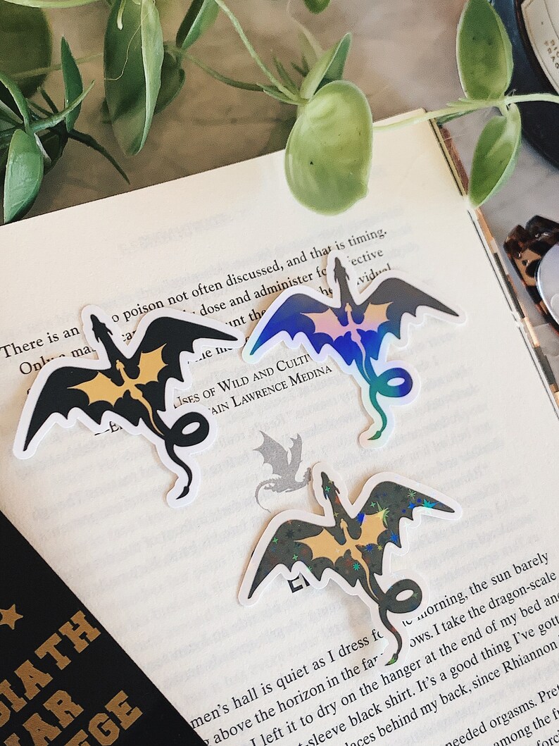 Pode incluir: Tr&ecirc;s adesivos de drag&atilde;o hologr&aacute;ficos com detalhes em preto e dourado. Os adesivos est&atilde;o em uma p&aacute;gina branca com texto sobre um livro.