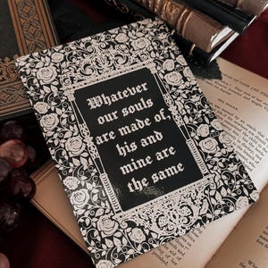 Könnte beinhalten: Eine schwarz-weiße dekorative Karte mit dem gotischen Zitat "Whatever our souls are made of, his and mine are the same". Die Karte hat einen floralen Rand und liegt auf einem offenen Buch und anderen antiken Büchern.
