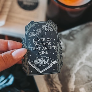May include: A black and white sticker featuring a book, a castle, and bats. The text on the sticker reads "Lover of Worlds That Aren't Mine".