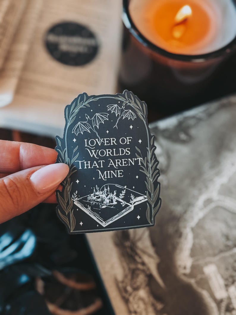 May include: A black and white sticker featuring a book, a castle, and bats. The text on the sticker reads "Lover of Worlds That Aren't Mine".