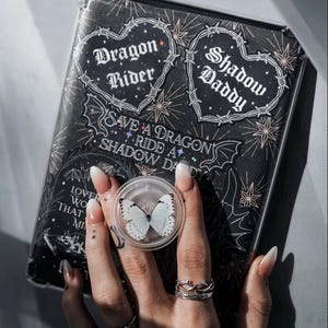 May include: A black phone case with a clear back, featuring a variety of stickers with gothic and edgy themes. The stickers include hearts with barbed wire, a dragon rider, and a shadow daddy. The case also has a sticker that says "Save a Dragon Ride a Shadow Da".