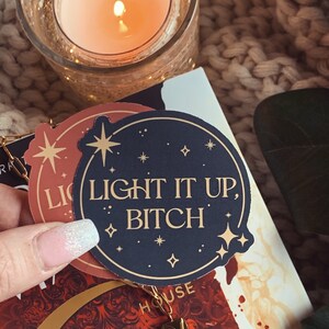 May include: A black and gold sticker with the text "Light it up, Bitch" surrounded by small gold stars. The sticker is on top of a book with a red and gold design.