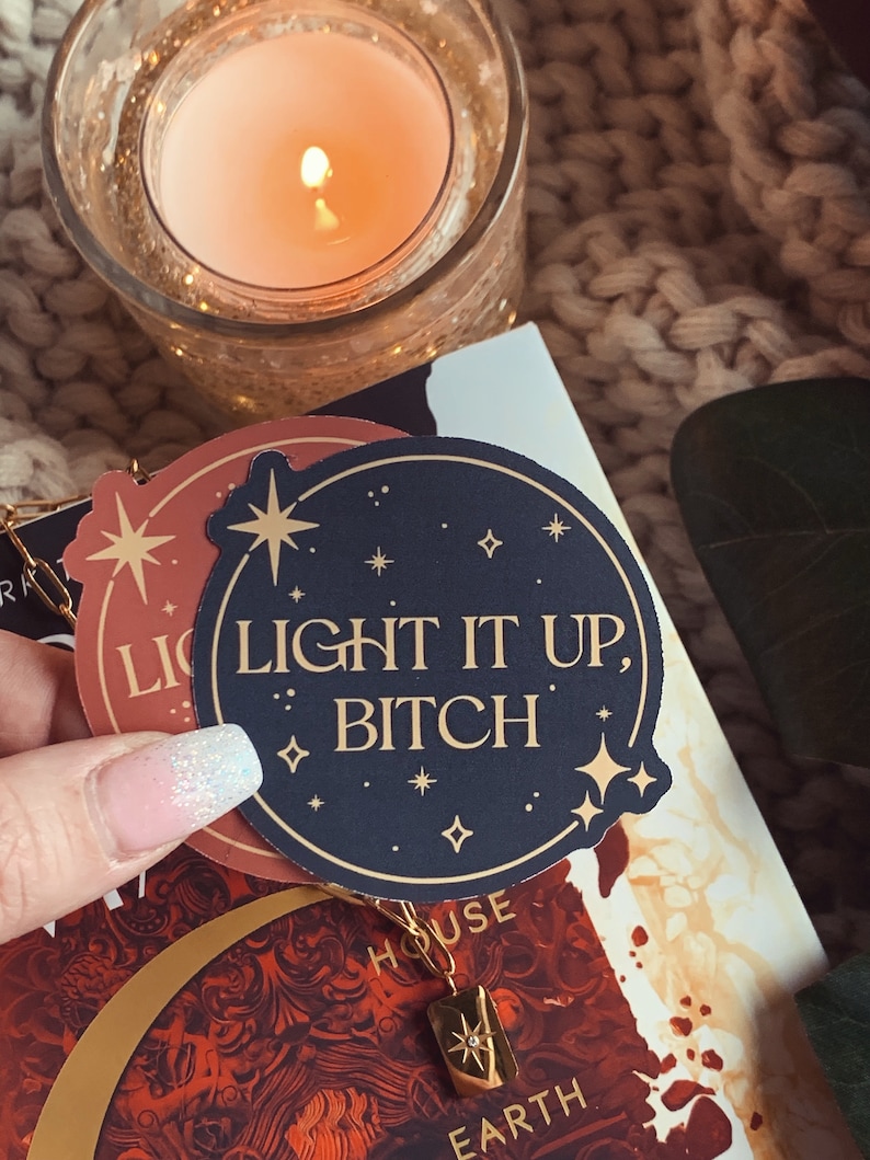 May include: A black and gold sticker with the text "Light it up, Bitch" surrounded by small gold stars. The sticker is on top of a book with a red and gold design.