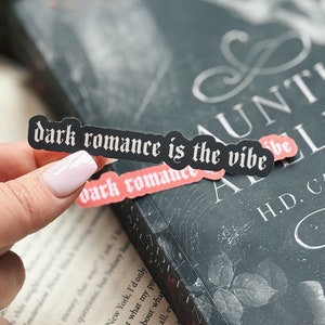 May include: A black and white sticker with the text "dark romance is the vibe" in a gothic font. The sticker is on a book cover with a black background and a white design.