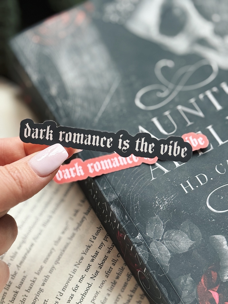 May include: A black and white sticker with the text "dark romance is the vibe" in a gothic font. The sticker is on a book cover with a black background and a white design.