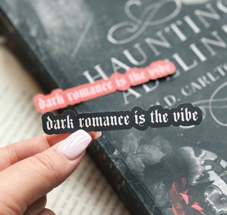 May include: Two black and white stickers with the text 'dark romance is the vibe' on a black book with white text that reads 'Haunting Addaline'.
