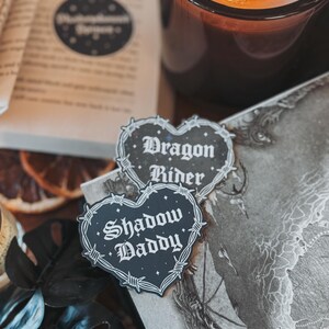 May include: Two black heart-shaped stickers with barbed wire borders. The top sticker says "Dragon Rider" and the bottom sticker says "Shadow Daddy".