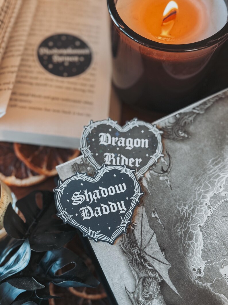 May include: Two black heart-shaped stickers with barbed wire borders. The top sticker says "Dragon Rider" and the bottom sticker says "Shadow Daddy".