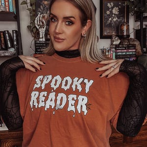 May include: A burnt orange t-shirt with the text "SPOOKY READER" in white dripping letters with a spider web design. The shirt has a black mesh sleeve.
