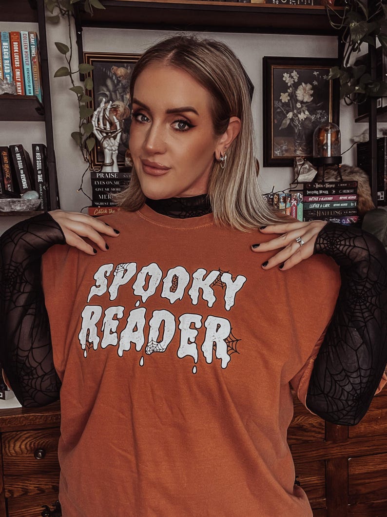 May include: A burnt orange t-shirt with the text "SPOOKY READER" in white dripping letters with a spider web design. The shirt has a black mesh sleeve.