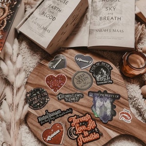May include: A wooden cutting board with a variety of stickers featuring characters and quotes from the Crescent City series by Sarah J. Maas. The stickers are in various colors and designs, including hearts, stars, and wings. The board is surrounded by books and a candle.