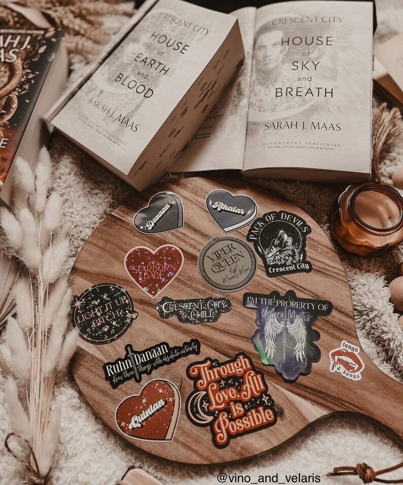 May include: A wooden cutting board with a variety of stickers featuring characters and quotes from the Crescent City series by Sarah J. Maas. The stickers are in various colors and designs, including hearts, stars, and wings. The board is surrounded by books and a candle.