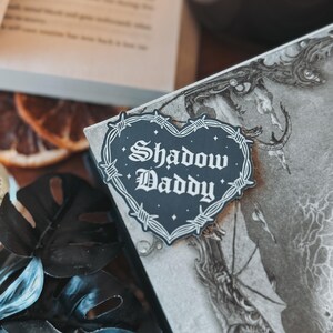 May include: A black and white heart-shaped sticker with barbed wire around the edge. The sticker says "Shadow Daddy" in a gothic font.