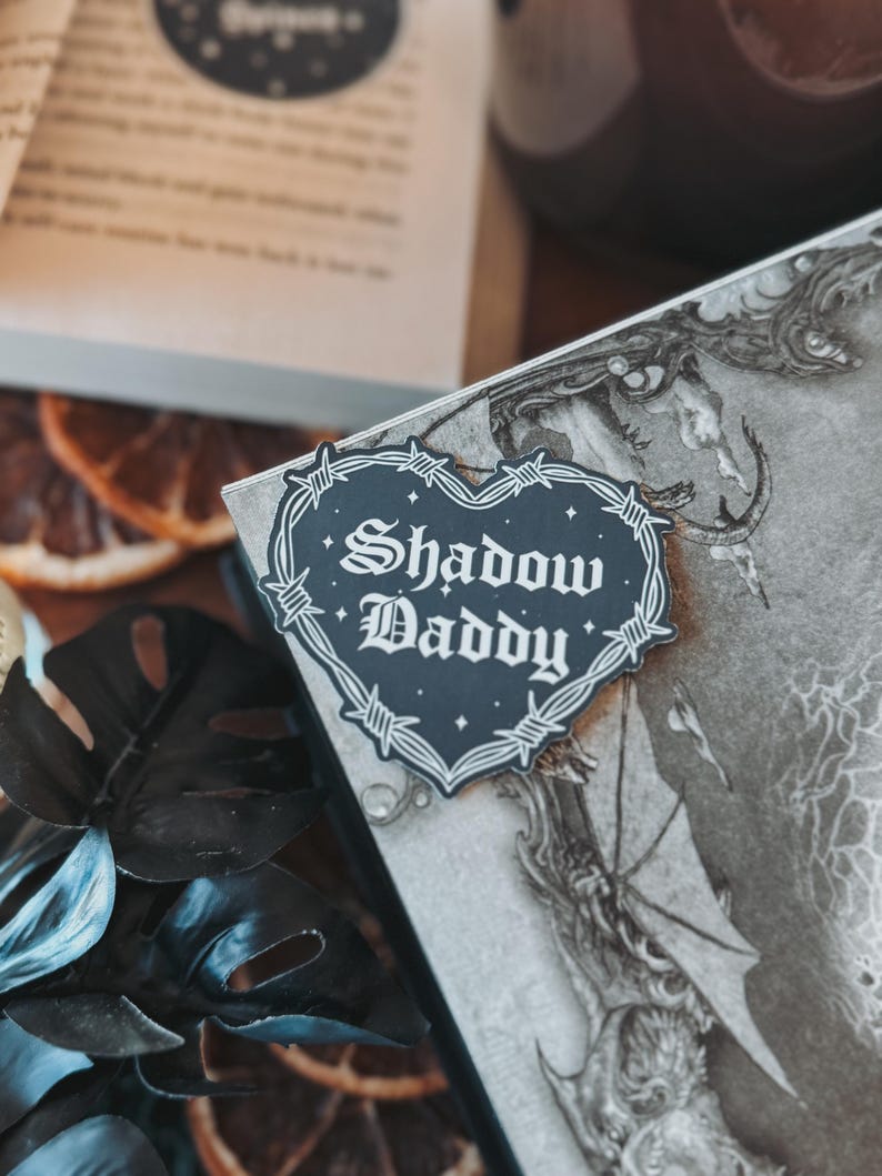May include: A black and white heart-shaped sticker with barbed wire around the edge. The sticker says "Shadow Daddy" in a gothic font.
