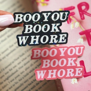 Puede incluir: Dos pegatinas negras y rosas con el texto "Boo You Book Whore" en un libro rosa y blanco. Las pegatinas tienen un acabado brillante.
