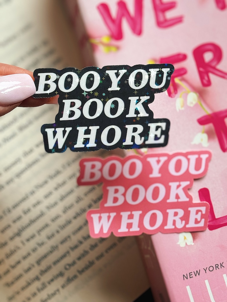 Puede incluir: Dos pegatinas negras y rosas con el texto "Boo You Book Whore" en un libro rosa y blanco. Las pegatinas tienen un acabado brillante.