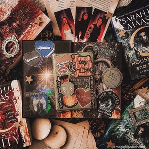 May include: A flat lay of various books and bookish items, including a book titled "House of Earth and Blood" by Sarah J. Maas, a book titled "Crescent City & Chill", and a book titled "Through Love All Is Possible". There are also several stickers, a small wooden box, and a star-shaped ornament.