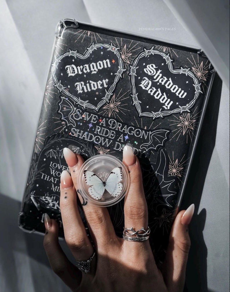 May include: A black phone case with a clear back, featuring a variety of stickers with gothic and edgy themes. The stickers include hearts with barbed wire, a dragon rider, and a shadow daddy. The case also has a sticker that says "Save a Dragon Ride a Shadow Da".