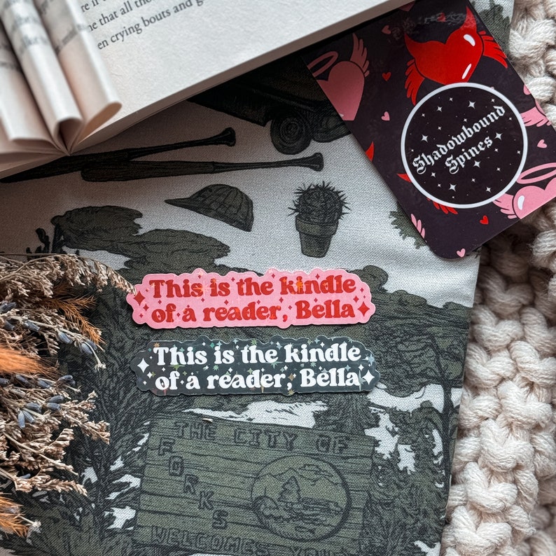 K&ouml;nnte beinhalten: Zwei rosa und schwarze Aufkleber mit dem Text "This is the kindle of a reader, Bella" auf einem wei&szlig;en und gr&uuml;nen Stoffhintergrund. Die Aufkleber haben eine glitzernde Oberfl&auml;che.