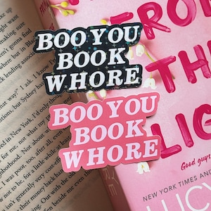 Puede incluir: Dos pegatinas con el texto "Boo You Book Whore" en letras negras y rosas con detalles de purpurina. Las pegatinas est&aacute;n colocadas en un libro rosa con el texto "We From The" visible.