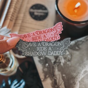 May include: Two stickers with a dragon design. The top sticker is pink with the text "Save a Dragon, Ride a Shadow Daddy". The bottom sticker is gray with the text "Save a Dragon, Ride a Shadow Daddy".