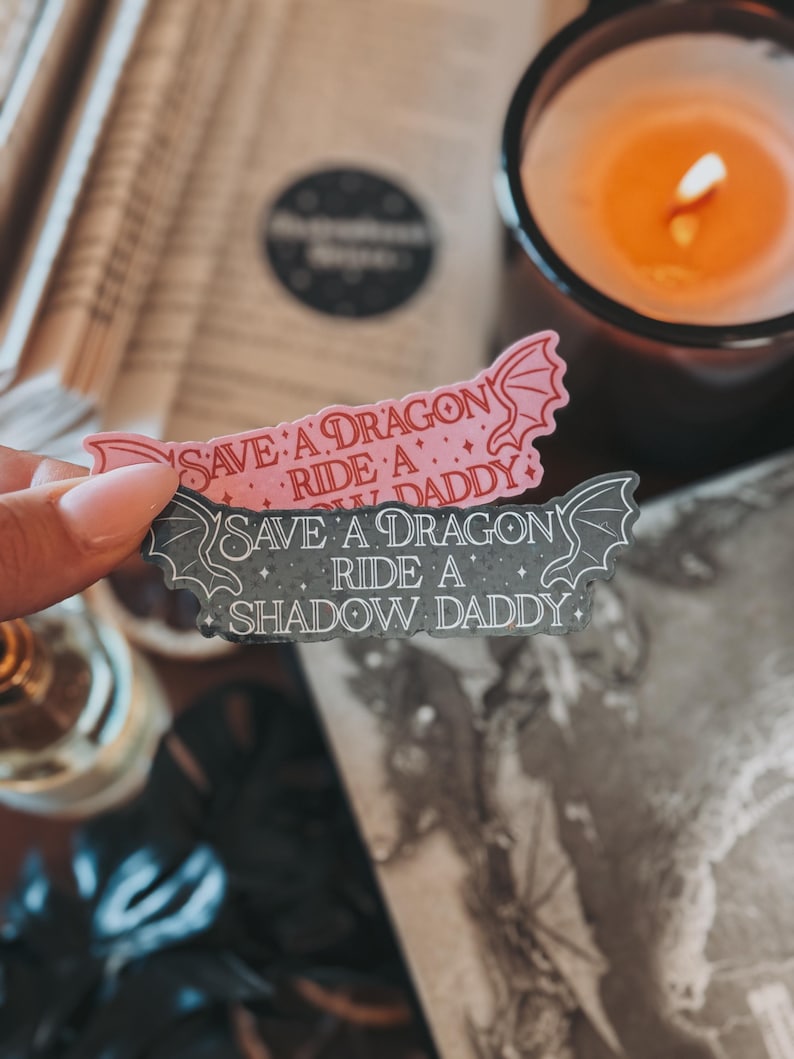 May include: Two stickers with a dragon design. The top sticker is pink with the text "Save a Dragon, Ride a Shadow Daddy". The bottom sticker is gray with the text "Save a Dragon, Ride a Shadow Daddy".