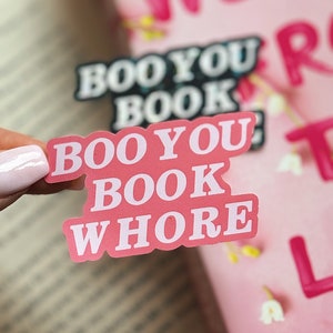 Puede incluir: Pegatina rosa con el texto "Boo You Book Whore" en letras blancas. La pegatina est&aacute; en un libro con una portada rosa.