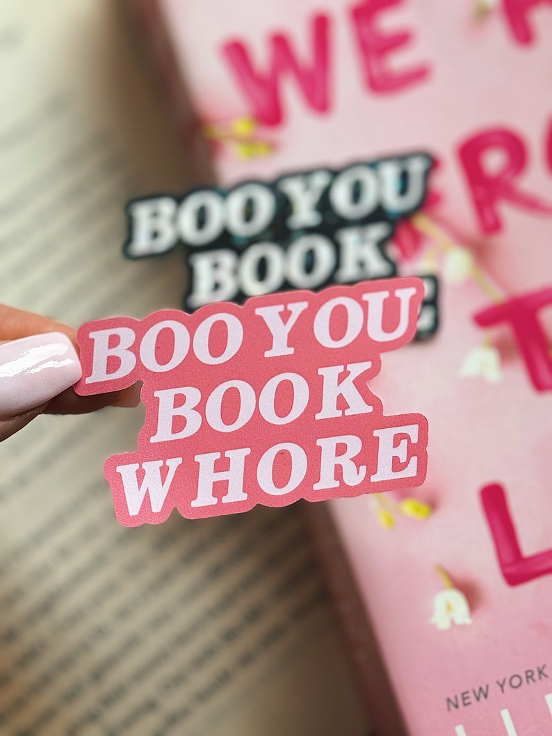 Puede incluir: Pegatina rosa con el texto "Boo You Book Whore" en letras blancas. La pegatina est&aacute; en un libro con una portada rosa.