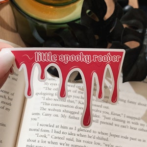 May include: A red bookmark with dripping blood and the text "little spooky reader" in white. The bookmark is on top of a book with text about a conversation.