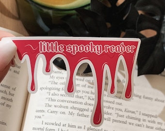May include: A red bookmark with dripping blood and the text "little spooky reader" in white. The bookmark is on top of a book with text about a conversation.