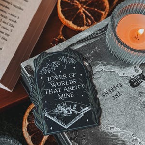 May include: A black and white sticker with a book open to reveal a small town with a starry sky. The text on the sticker reads "Lover of Worlds That Aren't Mine".