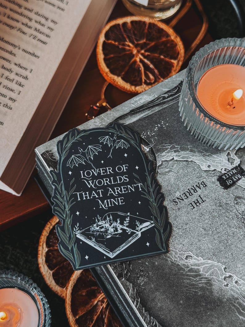May include: A black and white sticker featuring an open book revealing a small town under a starry sky. The text on the sticker reads "Lover of Worlds That Aren't Mine".