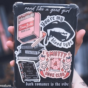 May include: A clear plastic case with various stickers on it. The stickers are pink, black, and red and feature book club themes, including the text "Slow Burn Book Club", "You're my blood type", and "Smutty Book Club".
