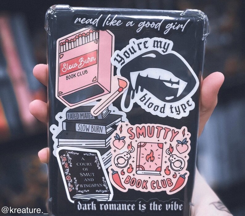 May include: A clear plastic case with various stickers on it. The stickers are pink, black, and red and feature book club themes, including the text "Slow Burn Book Club", "You're my blood type", and "Smutty Book Club".