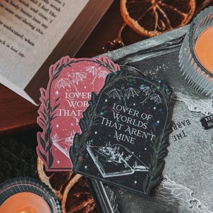 May include: Two black and pink stickers with a gothic aesthetic. The stickers feature a book with a forest scene and the text "Lover of Worlds That Aren't Mine".