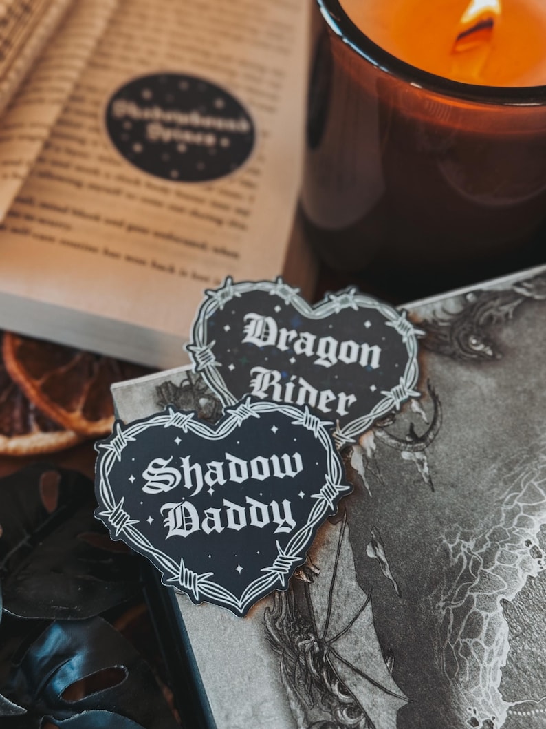 May include: Two black and white heart-shaped stickers with barbed wire borders. The stickers have white text that reads "Dragon Rider" and "Shadow Daddy".