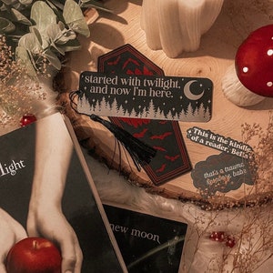 K&ouml;nnte beinhalten: Ein schwarzes und rotes Sarg-f&ouml;rmiges Lesezeichen mit einem silbernen Glitzerdesign und einem schwarzen Quasten. Das Lesezeichen zeigt den Text "started with twilight. + and now I'm here." und einen Halbmond.