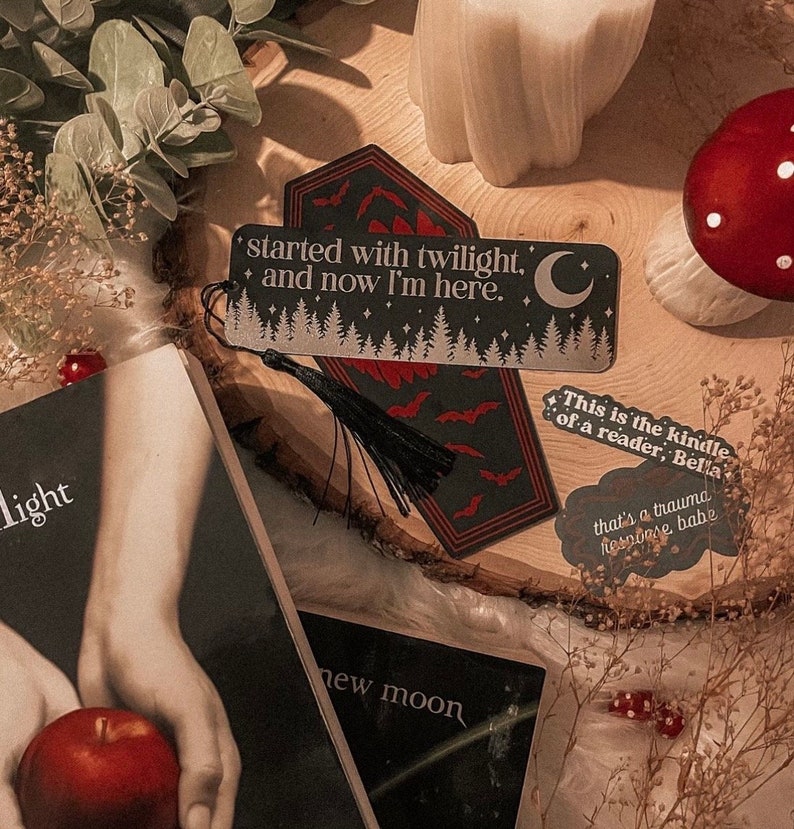 K&ouml;nnte beinhalten: Ein schwarzes und rotes Sarg-f&ouml;rmiges Lesezeichen mit einem silbernen Glitzerdesign und einem schwarzen Quasten. Das Lesezeichen zeigt den Text "started with twilight. + and now I'm here." und einen Halbmond.