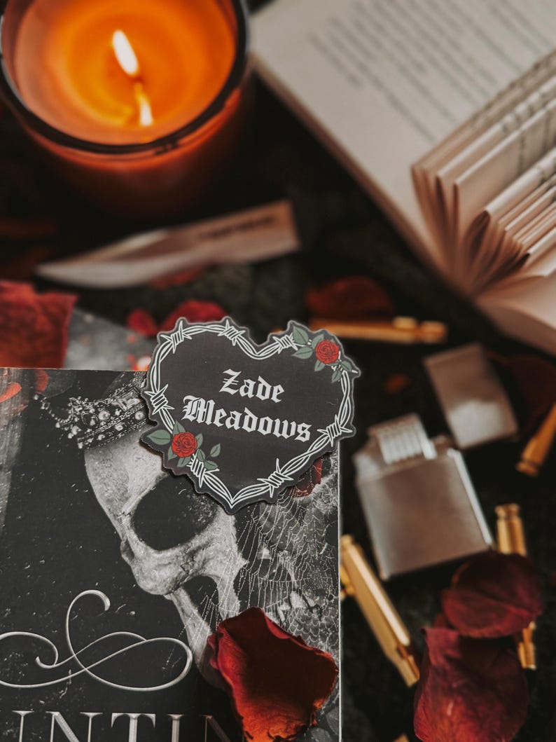 May include: A black and white book cover with a skull and a spiderweb design. A heart-shaped sticker with barbed wire and a rose is on the cover. The sticker says 'Zade Meadows'.