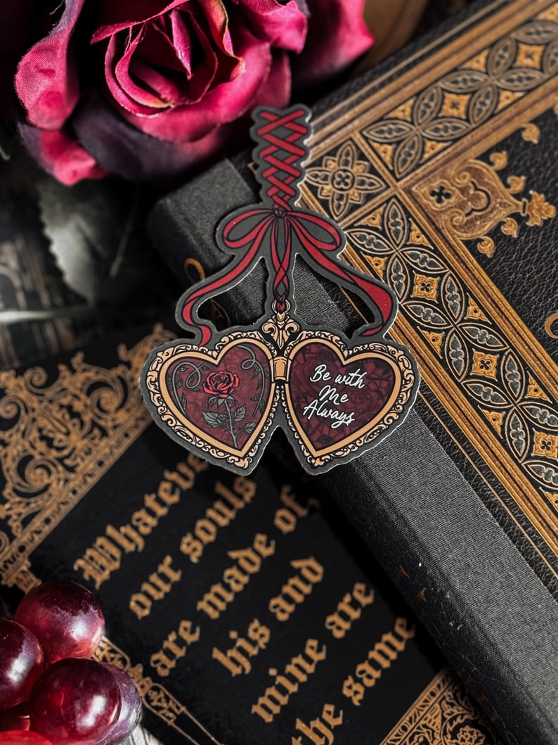 May include: A heart-shaped pendant with a red ribbon and the words "Be with Me Always." The pendant features a rose and ornate gold detailing. It rests on a dark book with gold lettering and a dark red rose in the background.