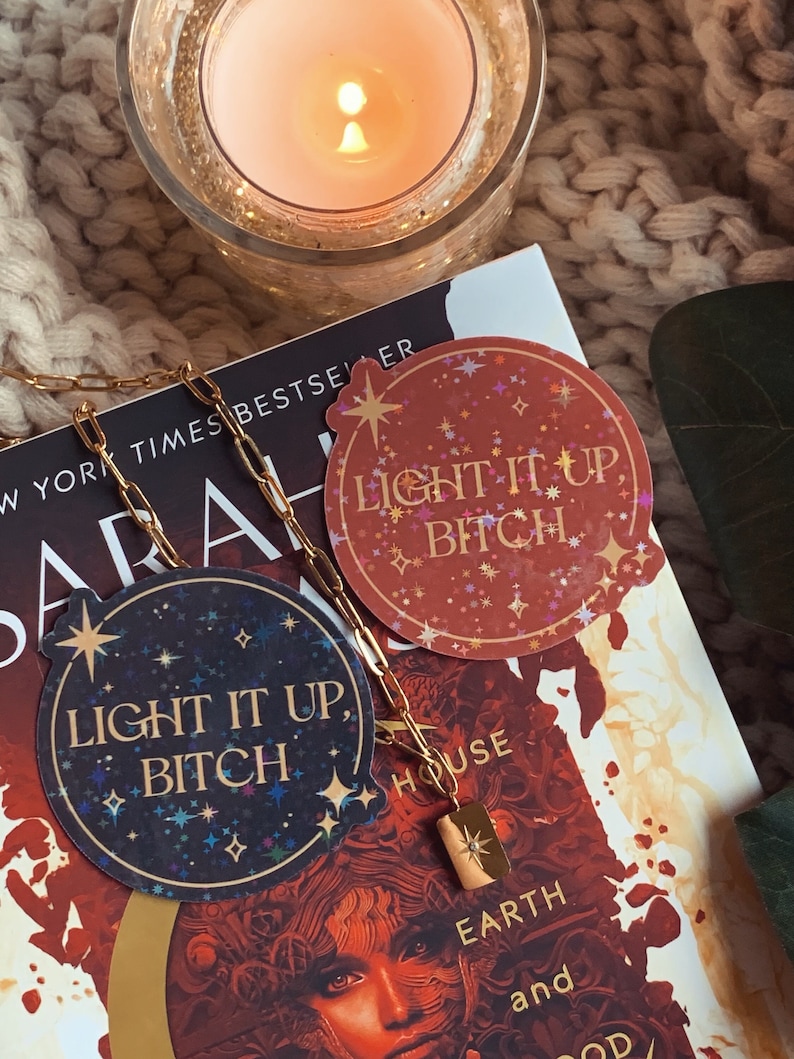 May include: Two holographic stickers with the text "Light it up, Bitch" in gold lettering on a dark blue and red background with gold stars. The stickers are on top of a book with the text "New York Times Bestseller" and "House of Earth and Blood".