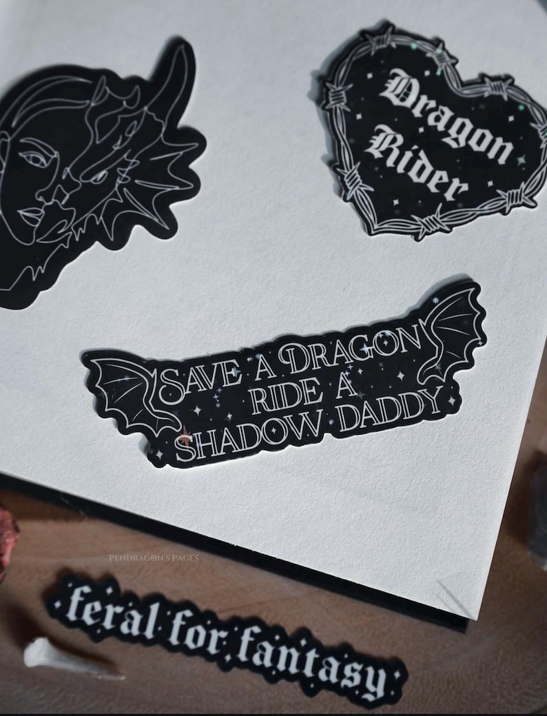 May include: Four black and white stickers with a fantasy theme. The stickers feature a dragon, a heart with barbed wire, a dragon with the text "Save a dragon, ride a shadow daddy", and the text "feral for fantasy".